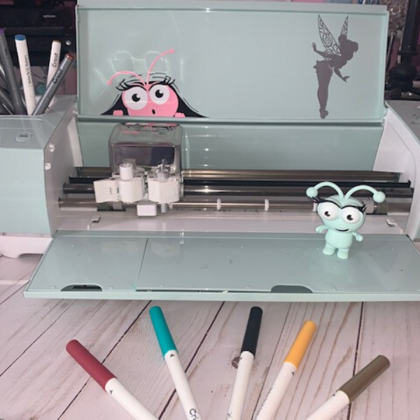 Open Cricut machine with little green Cricut standing on it and pens on display