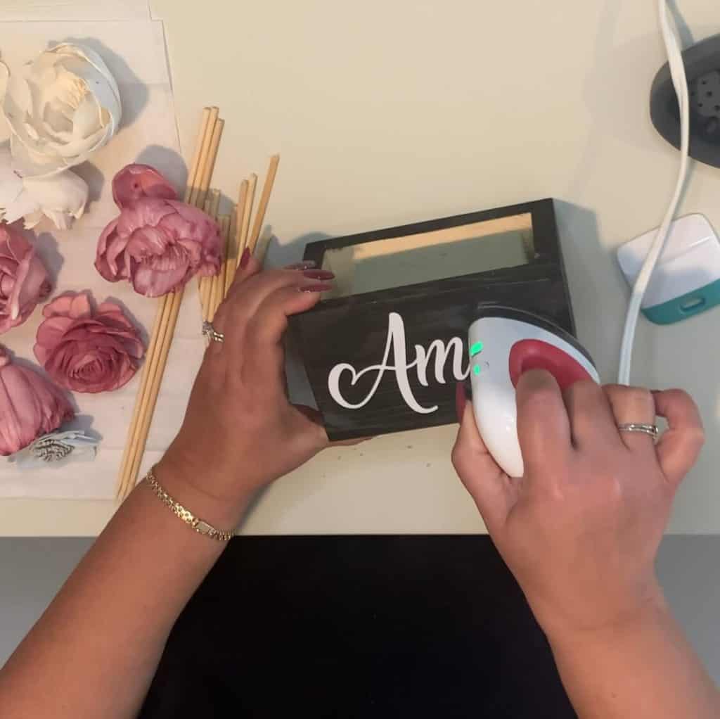 Woman using the Cricut mini easypress to iron on the word Amor to rectangular wooden box with merlot colored Sola wood flowers on the table